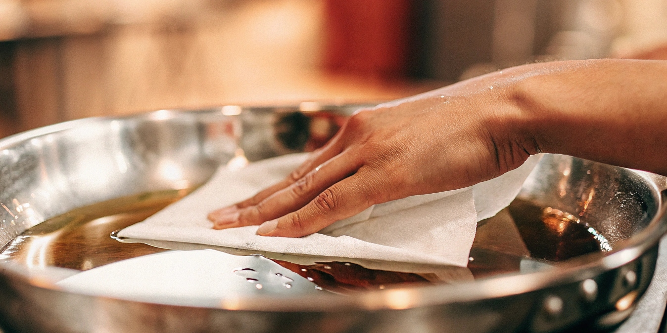 A hand wiping a thin layer of oil inside a titanium pan with a paper towel.